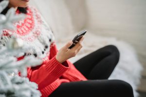Eine Person sitzt in einem warmen Norweger-Pullover und schwarzen Leggins auf einem Sofa und hält ein Telefon vor sich.