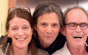 Zu sehen sind die Sprecher Katja Amberger, Wolfgang Hartmann und Martin Pfisterer. Alle drei lachen in die Kamera. Katja Amberger trägt eine Brille, die sie auf dem Foto ins lange braune Haar geschoben hat. Wolfgang Hartmann hat grau-braunes mittellanges Haar. Martin Pfisterer lacht mit geschlossenen Augen. Er trägt eine Brille und hat eine hohe Stirn und kurzes braun-graues Haar.