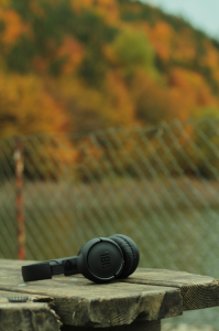 Ein schwarzer Kopfhörer liegt auf einer Holzoberfläche im Vordergrund. Im unscharfen Hintergrund ist ein Hügel mit Bäumen in leuchtenden Herbstfarben (Orange, Gelb, Grün) zu sehen.