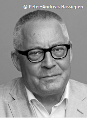 Portraitfoto Karl Schlögel: Er hat sehr kurze weiß-graue Haare, trägt eine schwarze Brille mit markantem Rand, ein weißes Hemd und ein helles Jacket. Er lächelt leicht ironisch in die Kamera.