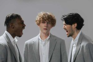 Zu sehen sind drei Männer, zwei davon schreien sich an, der dritte Mann steht dazwischen und sieht mit leerem Blick in die Kamera. Das ganze ist eine überzeichnete und damit ironische Darstellung eines Streits.